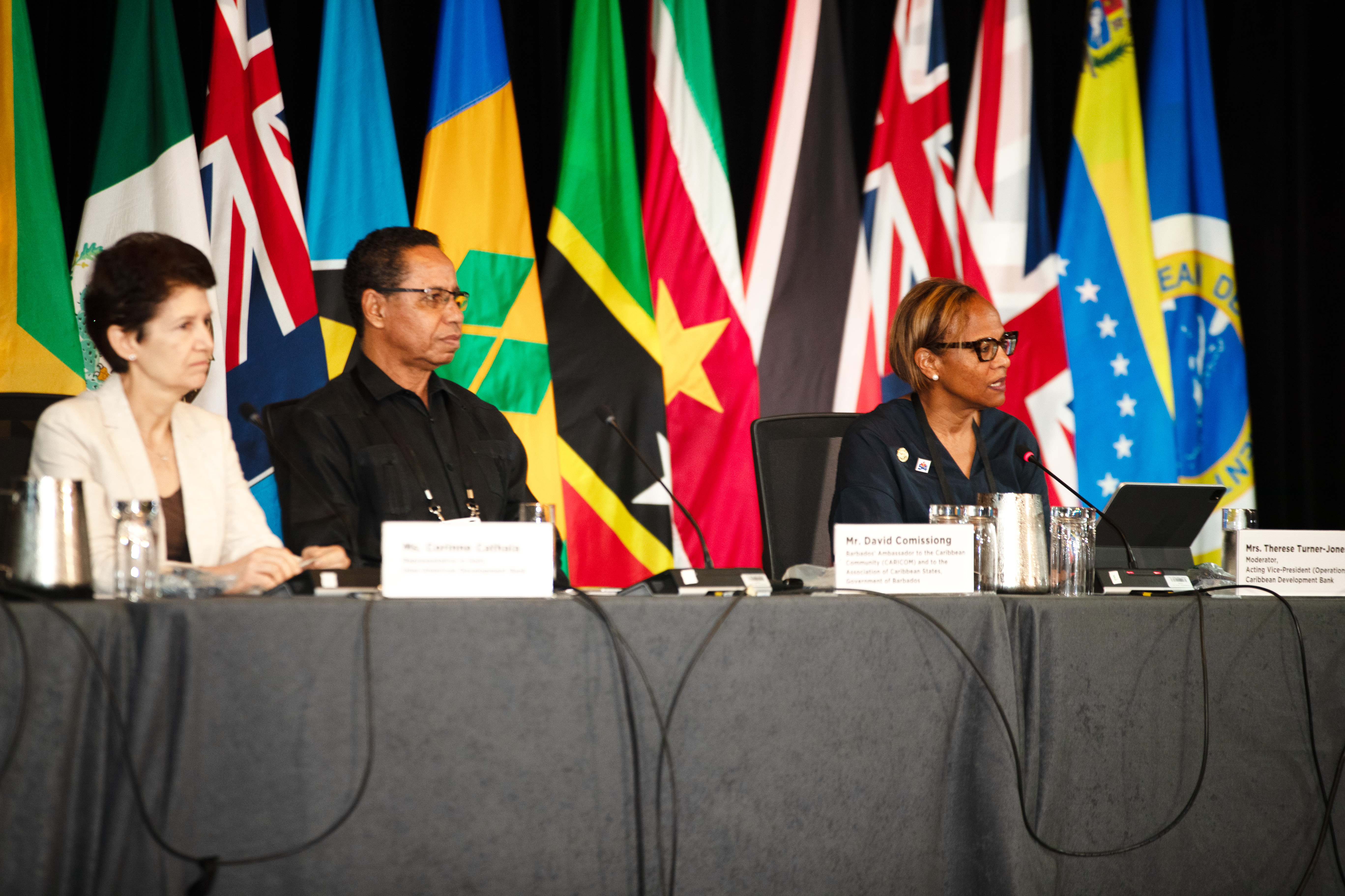 Three people sit at a table in front of several flags
