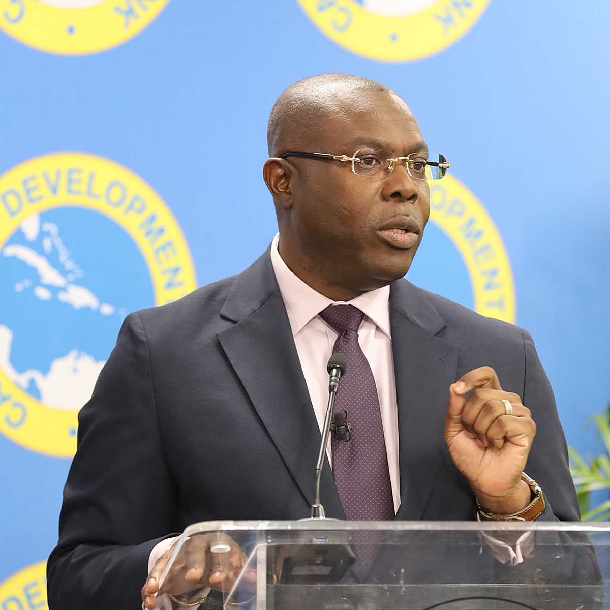 black gentleman in dark suit and tie with light coloured short, wearing glasses stands at lectern with left hand raised gesturing while speaking