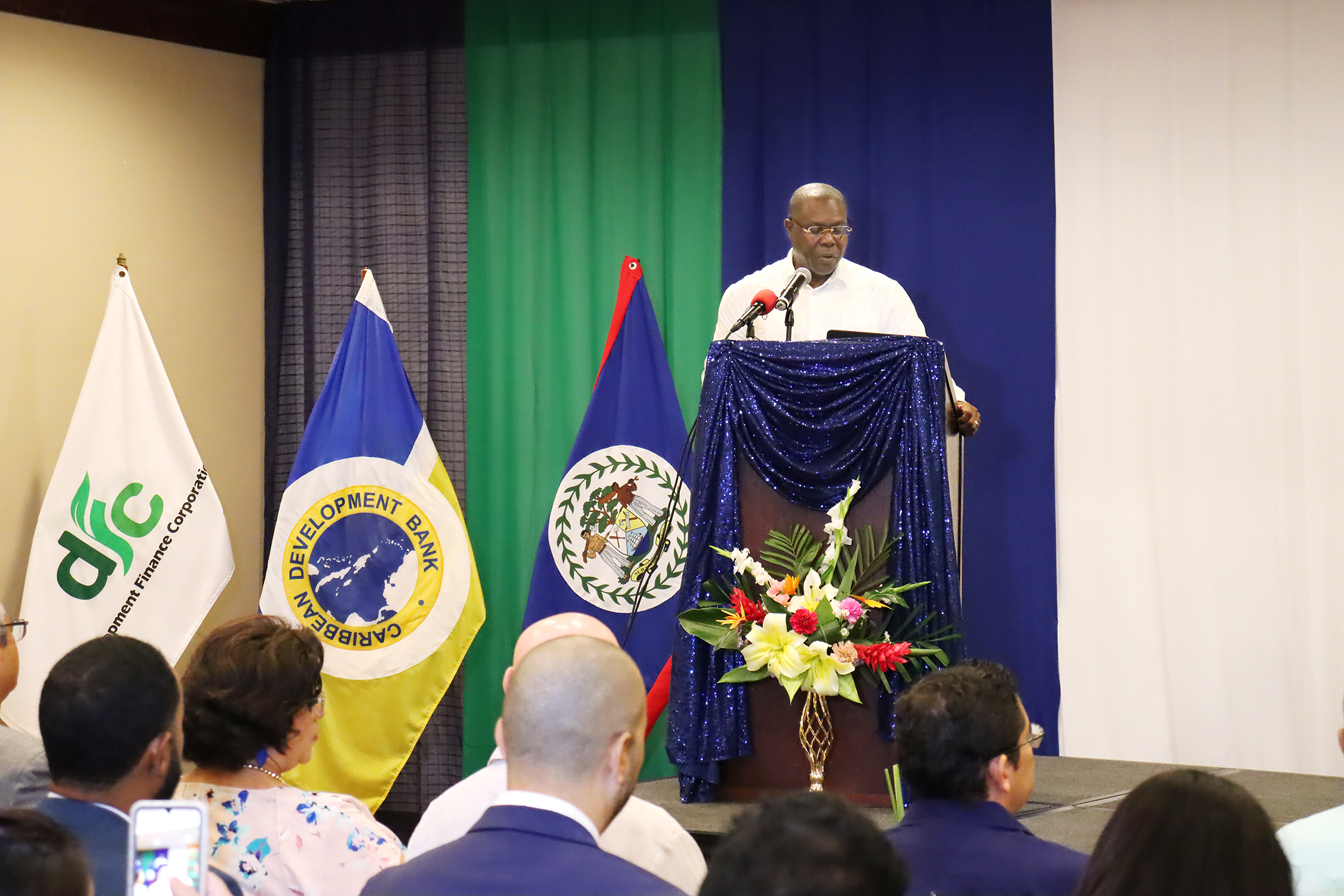 Black man standing at a lectern next to three flags delivering a speech