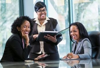 three black women in business attire in a boardroom. Two are seated and ne is standing the middle. They are smiling into the camera.