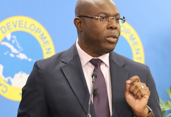 black gentleman in dark suit and tie with light coloured short, wearing glasses stands at lectern with left hand raised gesturing while speaking
